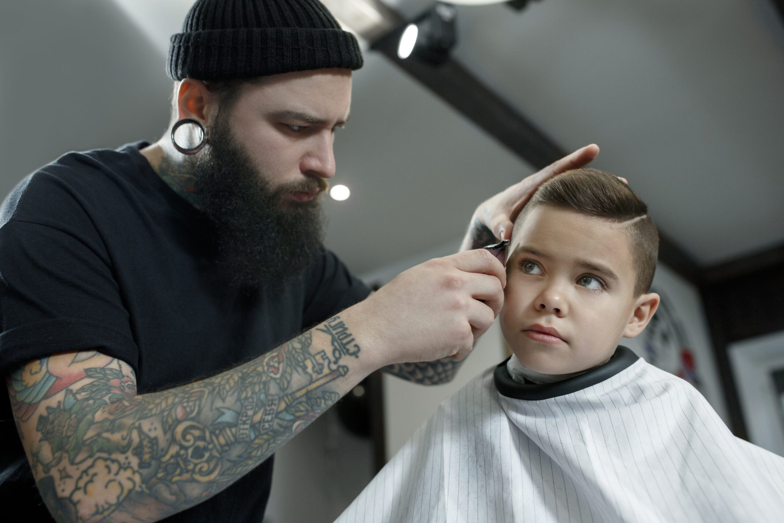 Children hairdresser cutting little boy against a dark background. Contented cute preschooler boy getting the haircut.The hand of the master have tattoo with the word shave